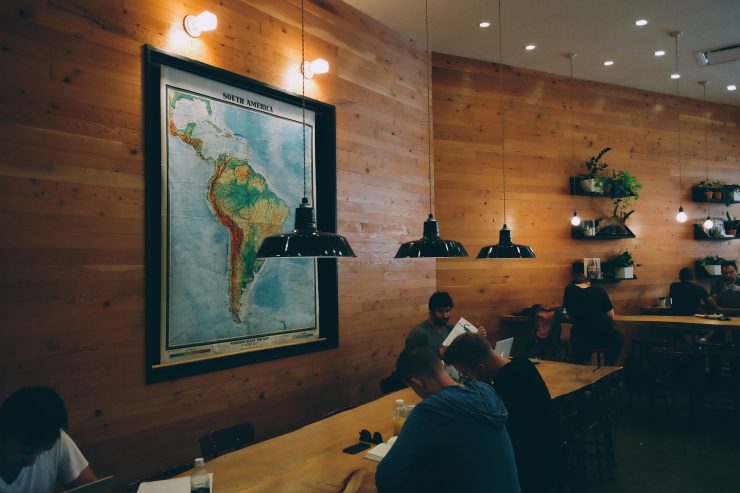 A foto mostra uma sala com várias pessoas estudando. Elas dividem uma longa mesa de madeira. Ao centro, um grande mapa com a foto da América Latina. 