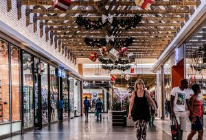 Imagem mostra o corredor de um shopping. As lojas estão decoradas para o período natalino. Vários consumidores passam com sacolas.