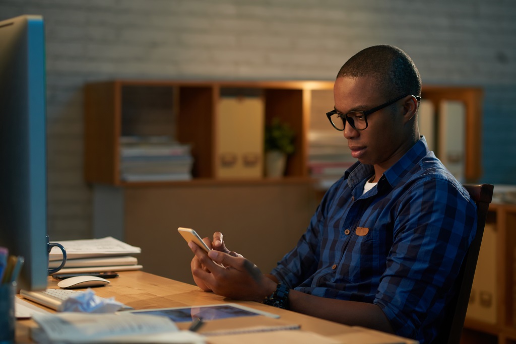 Imagem mostra um homem negro, trajando camisa azul, utilizando um celular. Ele usa óculos. Simboliza gestão em vendas.