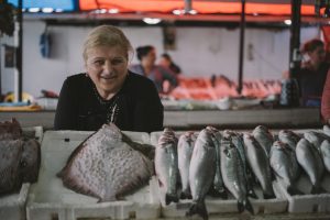 Microcrédito: na imagem, uma mulher idosa de cabelos loiros sorri para a câmera. Ela posa em frente a um balcão com vários peixes no que parece ser um mercado/feira livre.