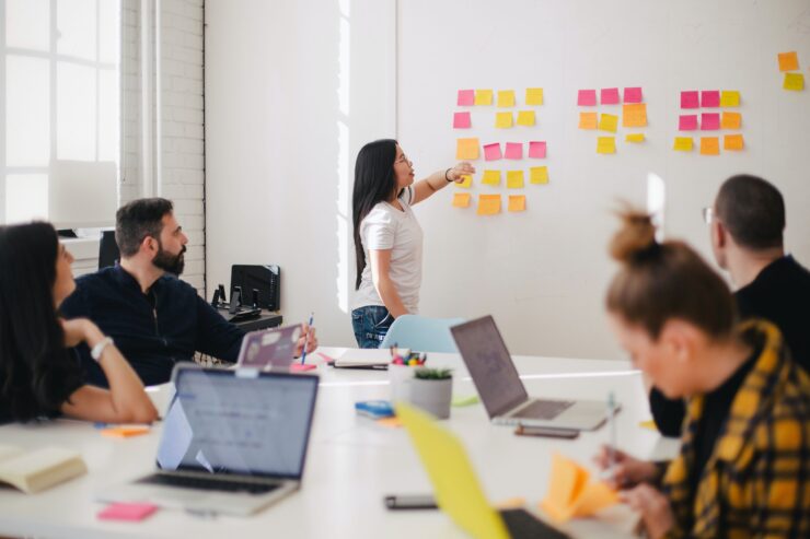 Uma mulher faz uma apresentação enquanto aponta para post-its. Uma mesa em frente dela, com várias pessoas e notebooks, é o centro da reunião.