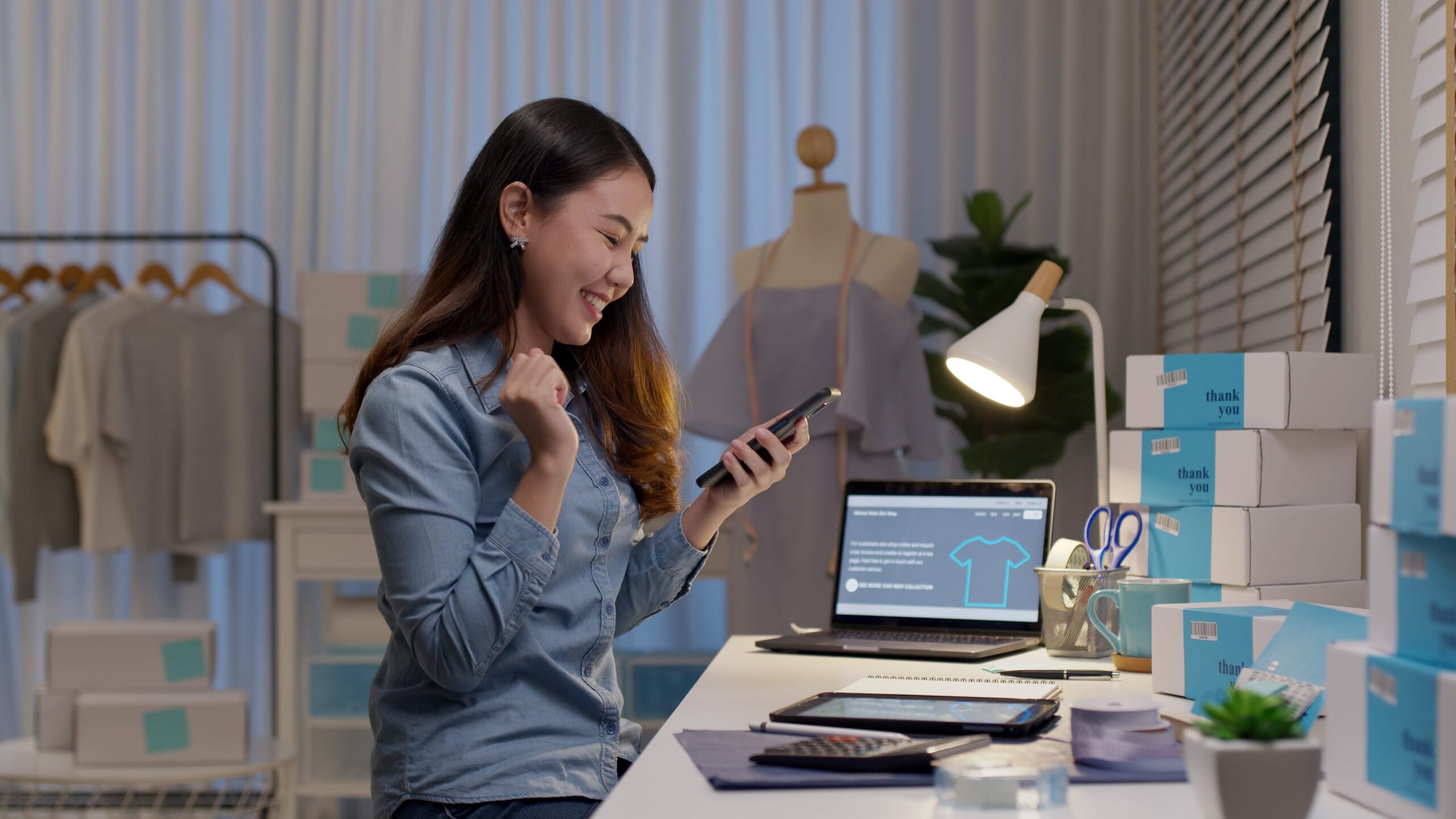 Transformação digital: uma mulher segura um celular enquanto sorri. Em frente a ela, há uma mesa com notebook aberto, caixas de envio, tablet e luminário. No fundo, há um manequim vestido.
