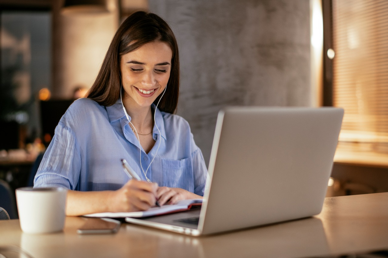 Mulher jovem e brnaca escreve sorrindo em frente a um notebook