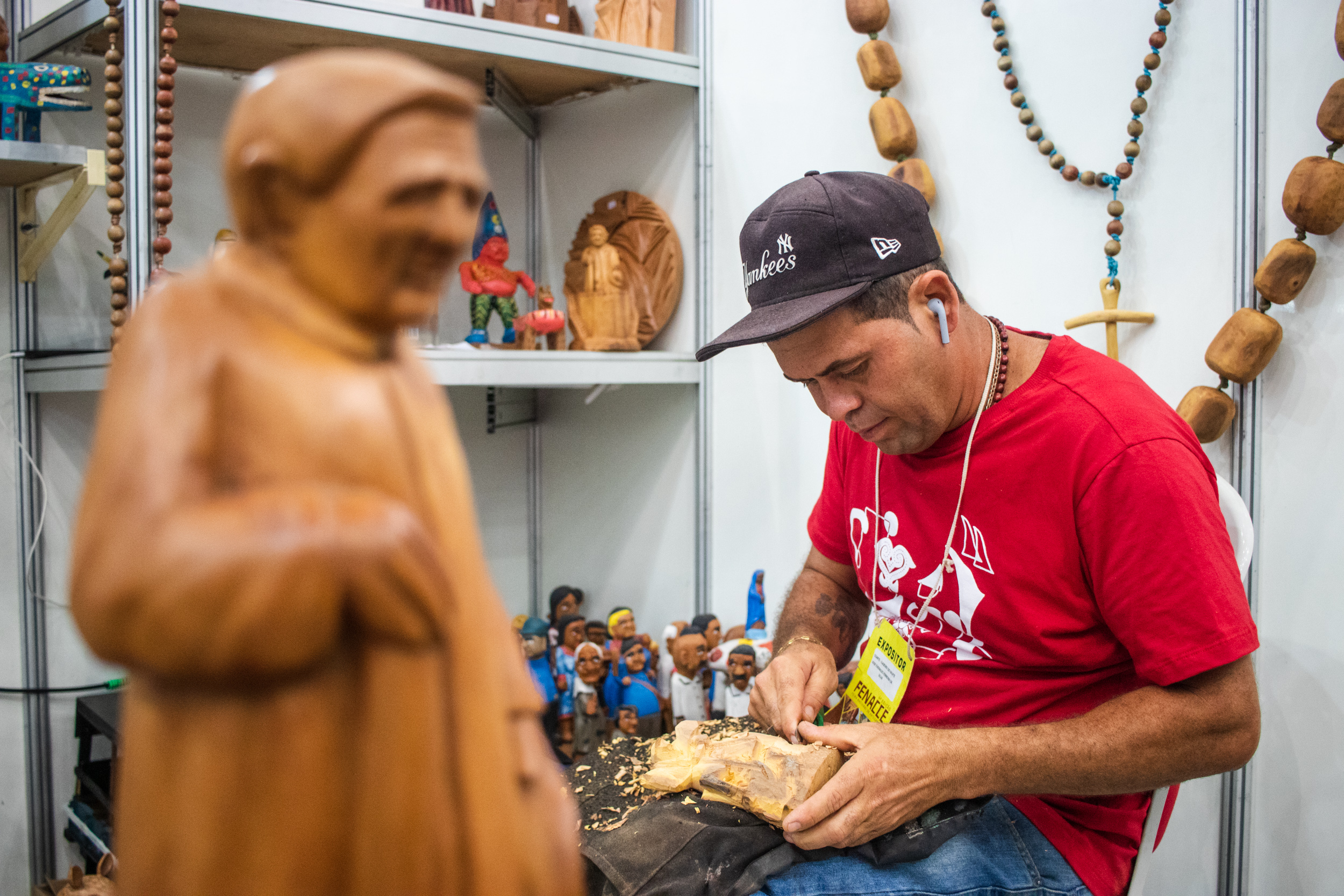 A foto mostra um artesão escupindo uma estátua de madeira em um estande cheio de outras esculturas. Ele usa boné, camisa vermelha, fones de ouvido e um crachá da Feira de Artesanato e Cultura do Ceará.
