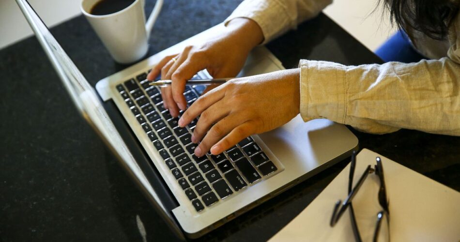 Mãos de uma pessoa branca digitando no teclado de um notebook. Entre os dedos de uma das mãos está uma caneta. Uma xícara de café está ao lado do computador.