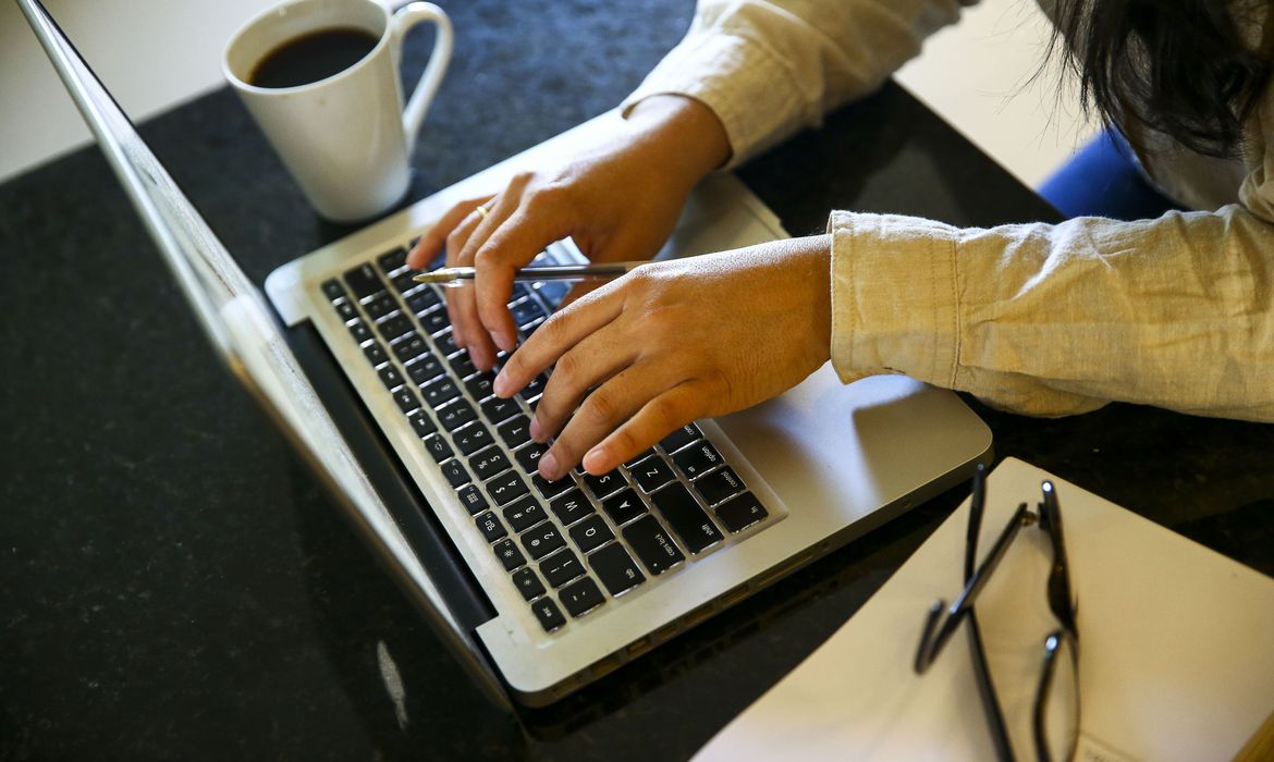 Mãos de uma pessoa branca digitando no teclado de um notebook. Entre os dedos de uma das mãos está uma caneta. Uma xícara de café está ao lado do computador. 