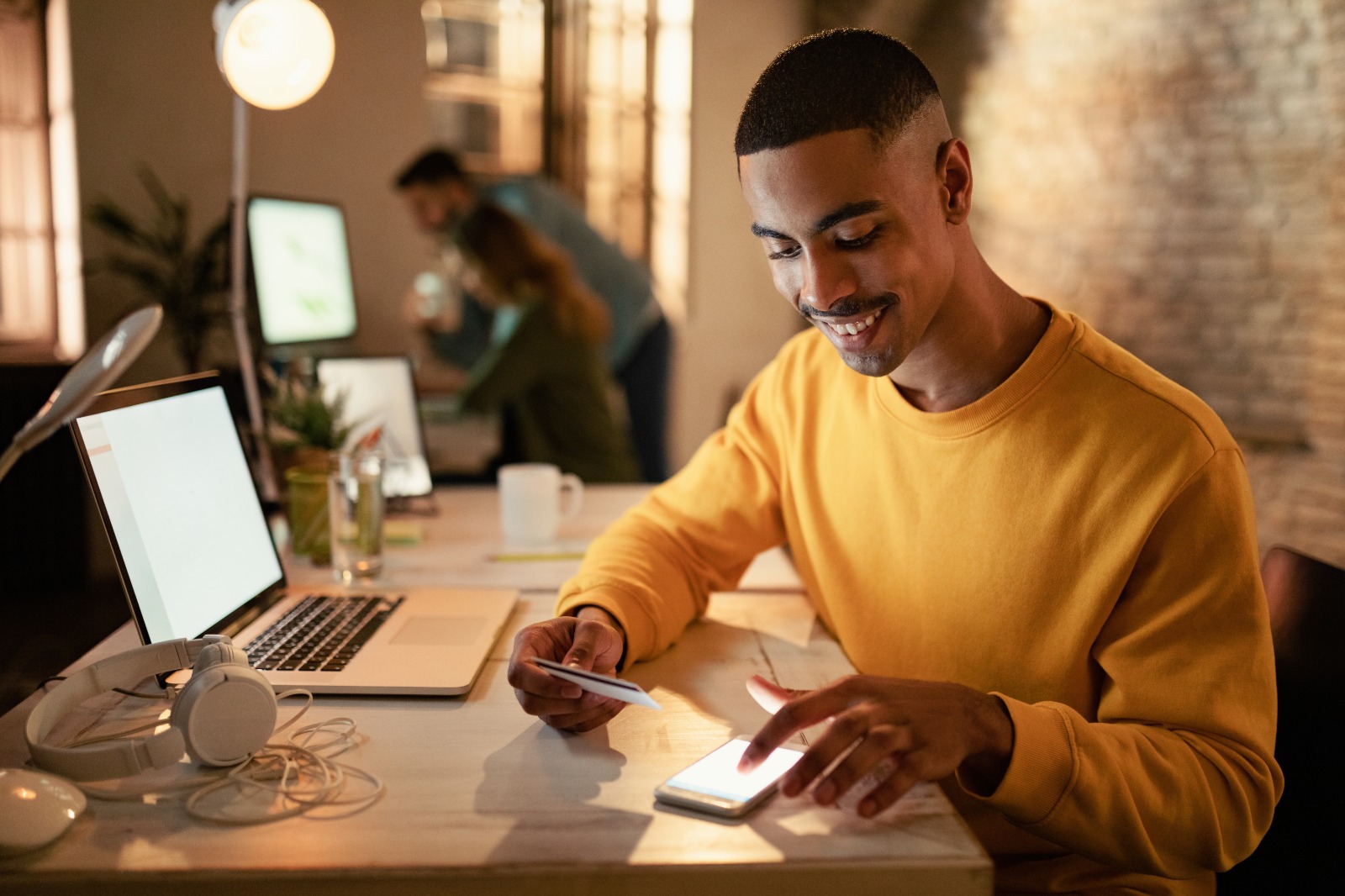 Um homem jovem negro sorri enquanto digita no celular. Ele está sentado em frente a uma mesa, em que há um computador e um pequeno vaso de plantas.