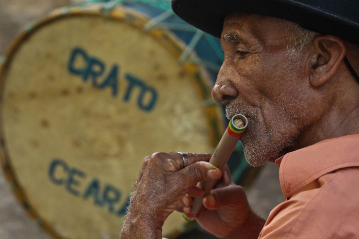 Homem idoso toca uma flauta de madeira. Ao fundo, um tambor com os dizeres "Crato" e "Ceará".