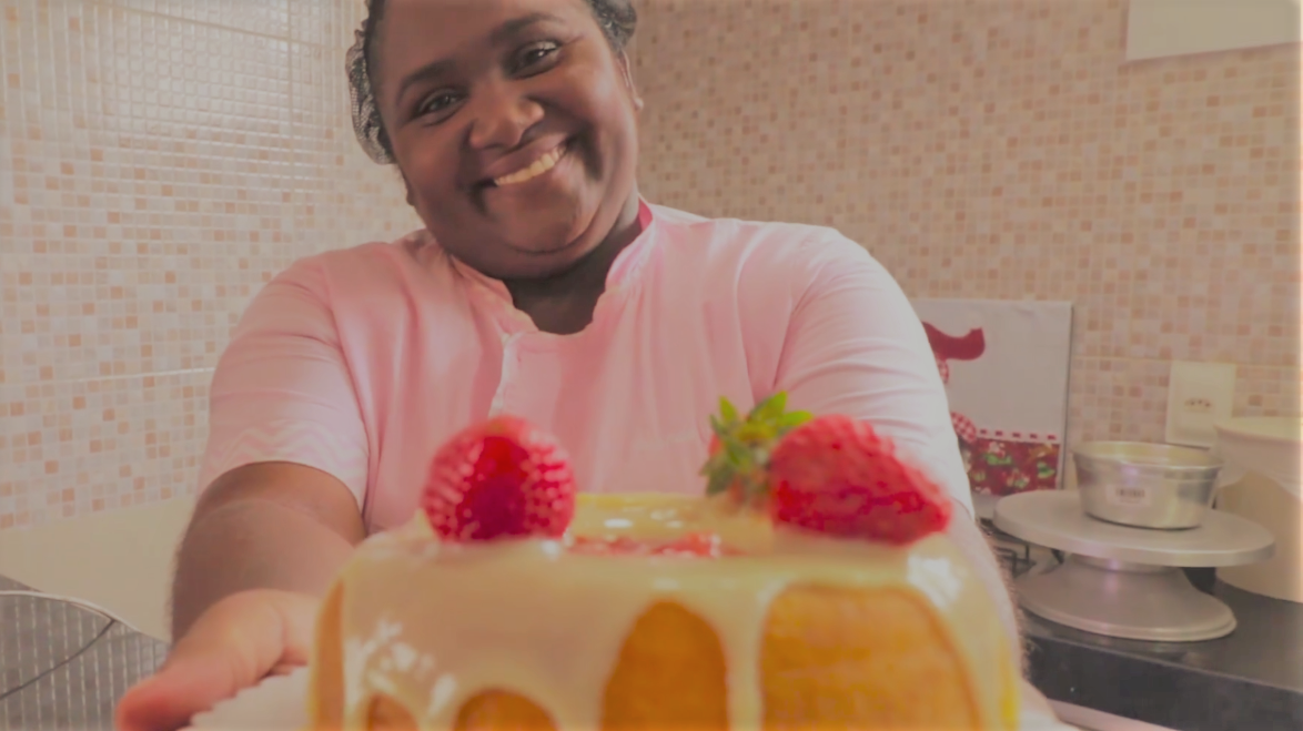 Iolanda Galvão segura um bolo confeitado com morangos e sorri para a câmera. Ela é uma mulher negra, com roupa rosa e traja uma touca na cabeça.