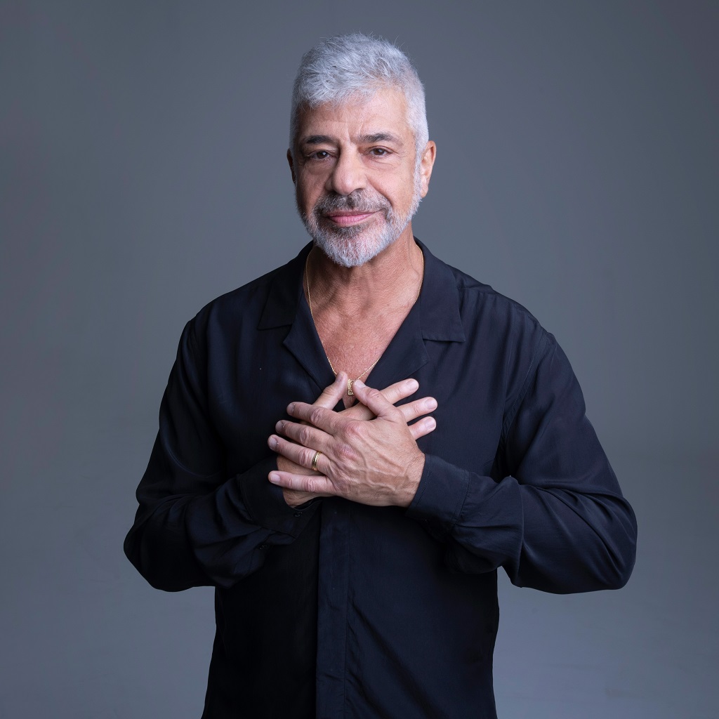 Foto do cantor Lulu Santos. Ele usa uma blusa preta e faz um sinal sobre o coração.
