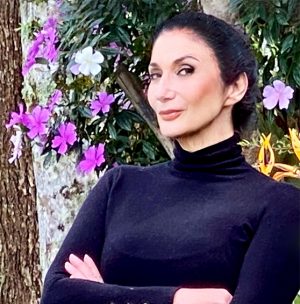 Na foto, Zizi Possi usa uma blusa preta e justa de gola alta, está com os braços cruzados e o cabelo escuro preso em um coque. Ela sorri para a câmera e está em frente a um muro com flores roxas.