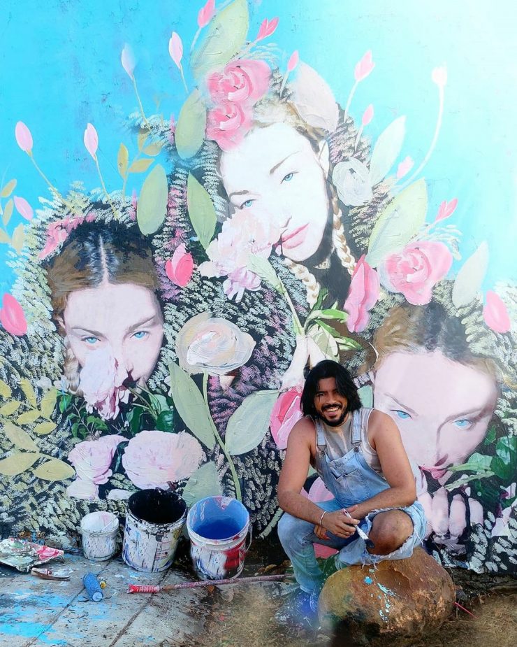 Artista Wanderson Petrova sentado em frente a um muro grafitado por ele com imagem da Madona; obras dele são exemplos de arte urbana