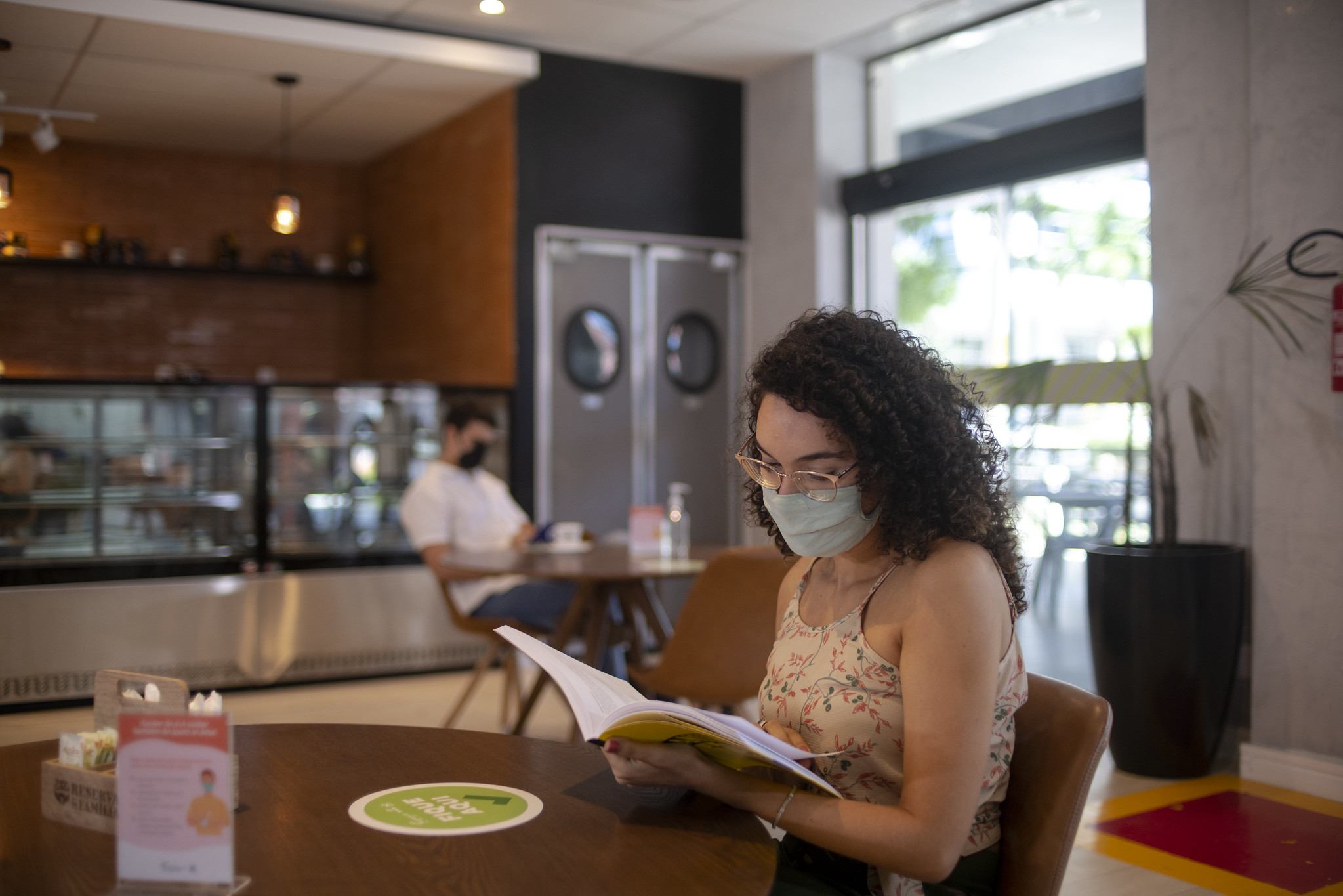 Livraria Senac Ceará: na imagem, uma mulher branca com cabelo cacheado castanho folheia um livro em uma mesa. Ela usa óculos e máscara de proteção.