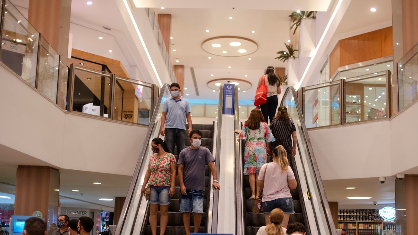 Expectativas para 2021: na imagem, duas escadas rolantes de um shopping estão cheias de clientes subindo (à direita) e descendo (à esquerda). Os clientes usam máscara e usam roupas coloridas.