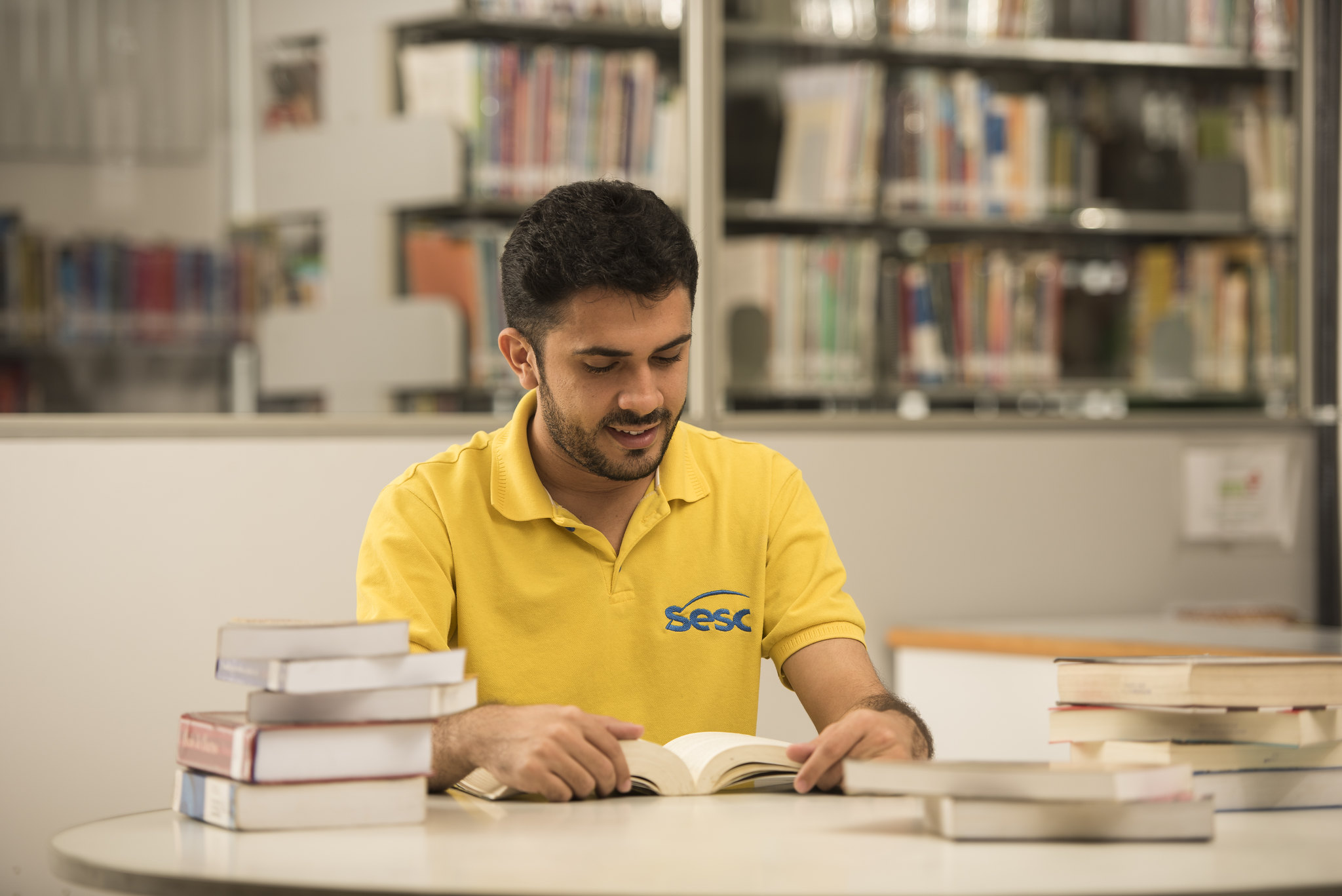 Na imagem, um homem está sentado em frente a uma mesa cheia de livros, lendo um exemplar. Ele possui cabelo e barba curtos e pretos e usa uma camisa amarela onde se lê o nome "Sesc", escrito em azul.