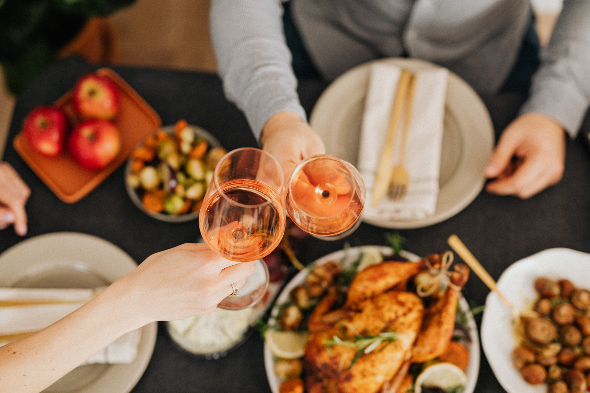 Ceias de Natal: a imagem mostra duas mãos brindando taças de vinho rosé. Embaixo há uma mesa onde se pode ver um peru e alguns outros pratos em segundo plano.