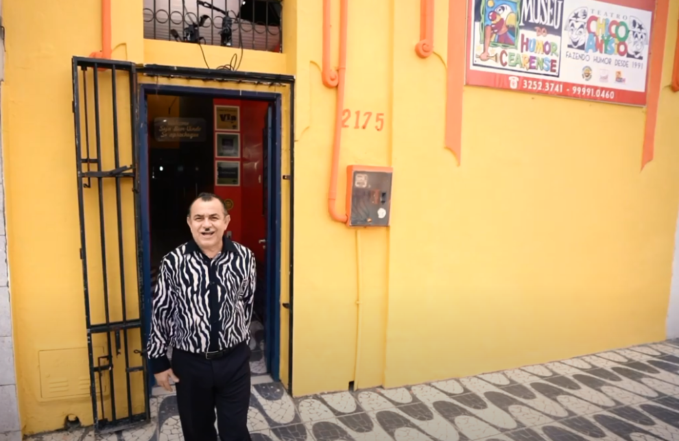 Na imagem, um homem está em frente ao Museu do Humor Cearense, um imóvel amarelo e laranja com detalhes pretos.