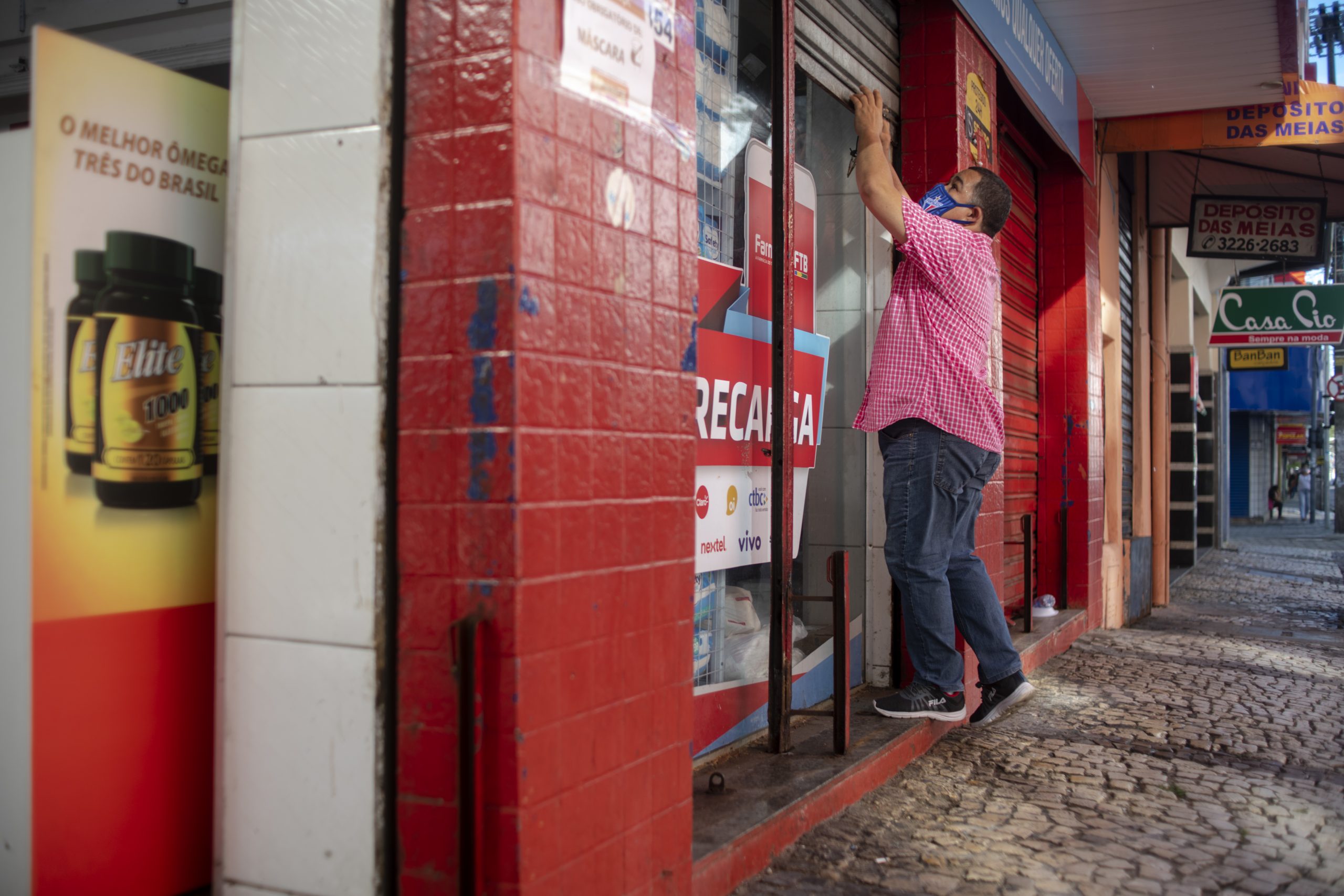 Lockdown: a imagem mostra uma loja de paredes vermelhas sendo fechada por um homem de máscara. Ele usa camisa de cor rosa, calça jeans e tênis.