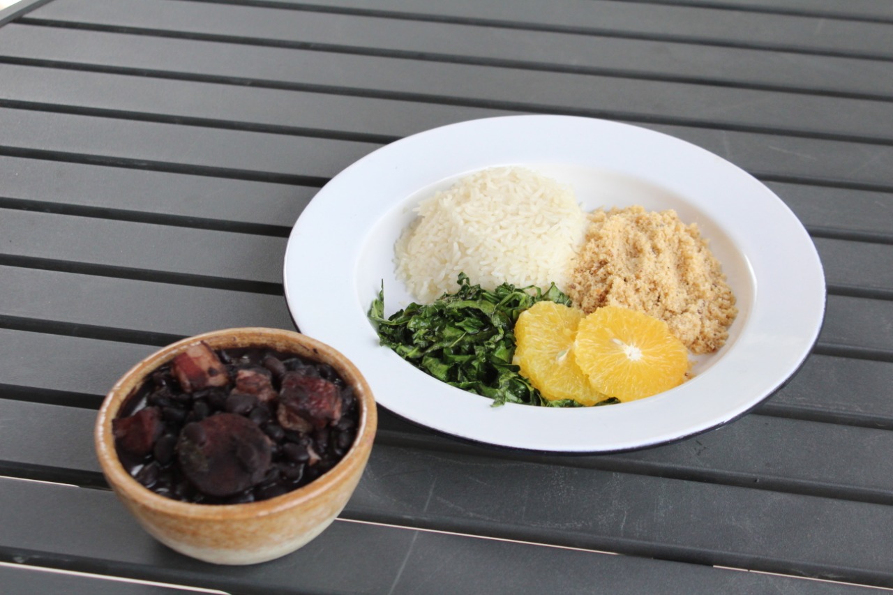 Delivery Café Senac: na imagem, há dois pratos: uma cumbuca com feijoada e um prato branco fundo com arroz, farofa, couve e rodelas de laranja. Os pratos estão em cima de uma mesa de madeira escura.