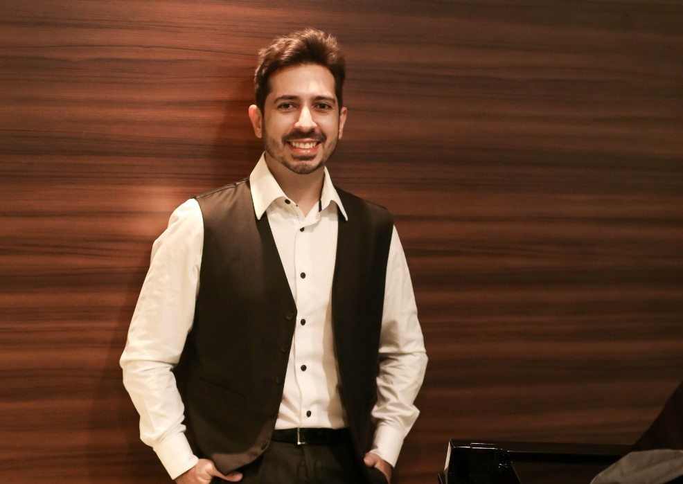 Dia da Mulher: pianista Felipe Adjafre posa ao lado de um piano. Ele usa uma camisa social branca, calça e colete pretos e sorri para a câmera.