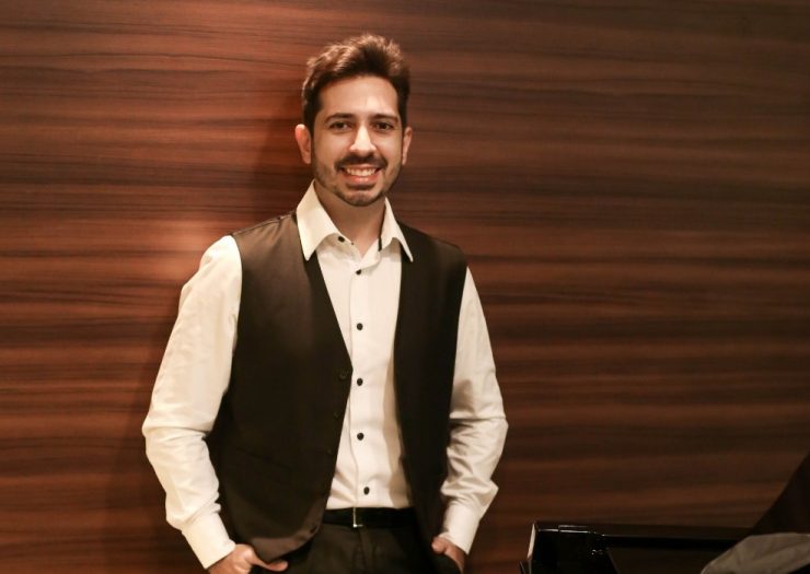 Dia da Mulher: pianista Felipe Adjafre posa ao lado de um piano. Ele usa uma camisa social branca, calça e colete pretos e sorri para a câmera.