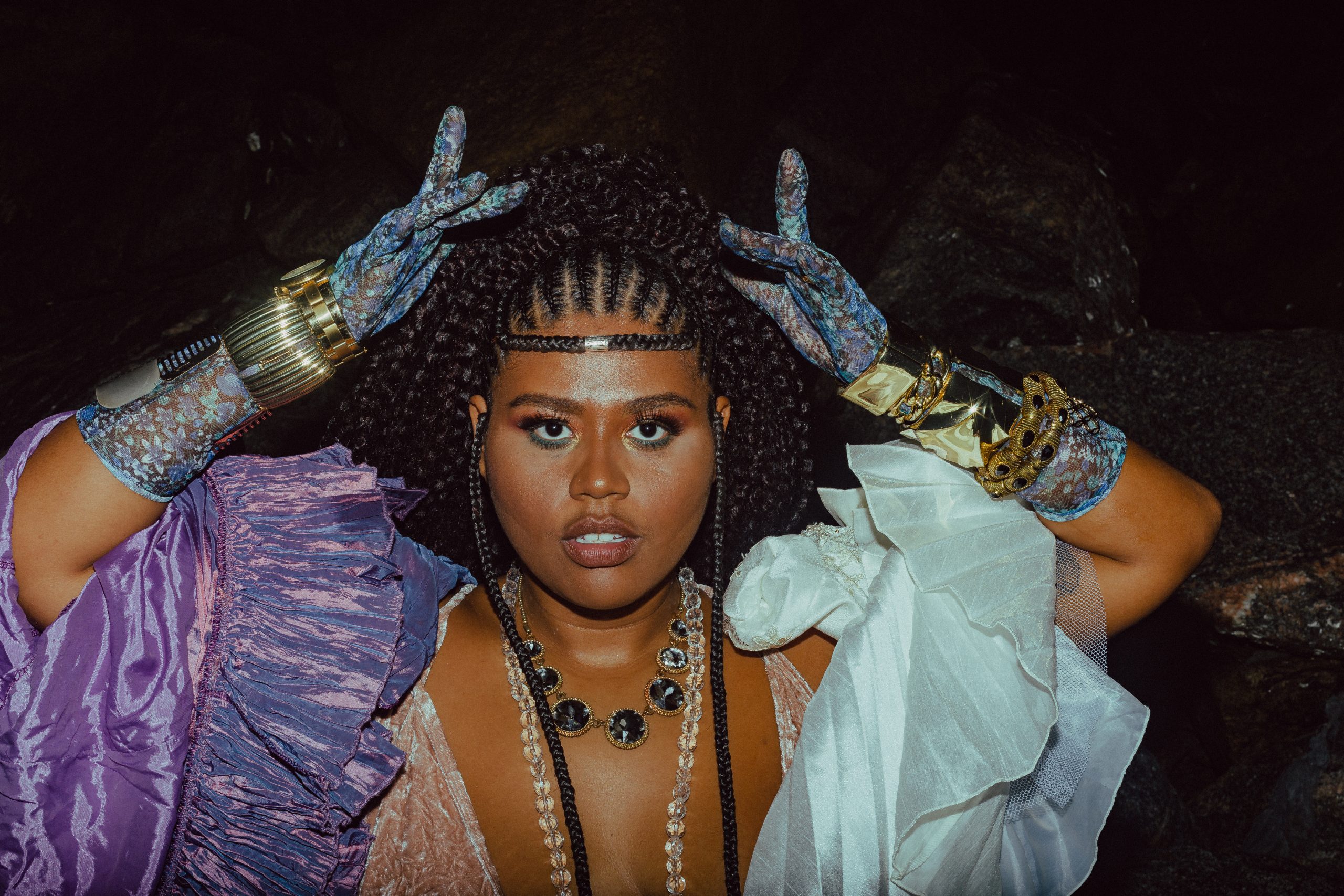Programação online Fecomércio: na imagem, a cantora Luiza Nobel olha para a foto com as mãos próximas à cabeça. Ela tem a pele negra e o cabelo preto com tranças, está com uma maquiagem artística, luvas azuis e um vestido azul com violeta.