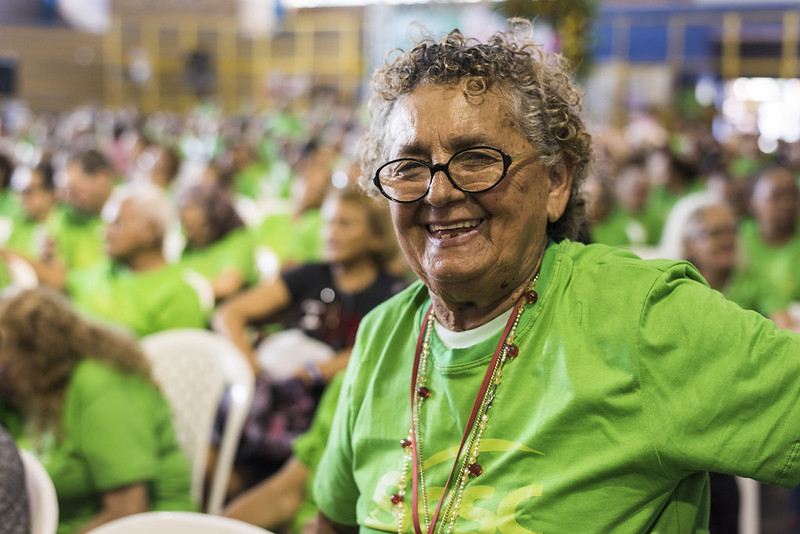 Envelhecimento ativo: na imagem, uma mulher idosa sorri para a câmera. Ela usa camisa verde, óculos com armação preta e cabelos encaracolados e grisalhos curtos.