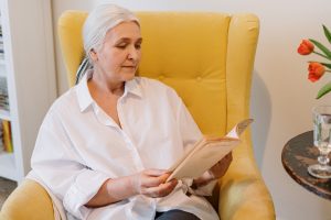 Dia das Mães: na imagem, uma mulher lê um livro em uma poltrona amarela. Ela tem cabelos brancos e lisos, pele branca e usa uma camisa social de cor branca.