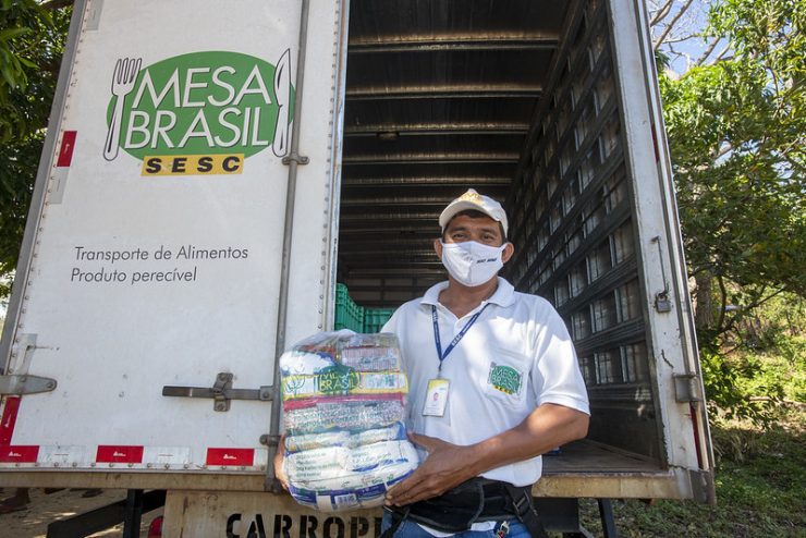 Mesa Brasil Sesc: na imagem, um homem de máscara, boné e camisa brancos segura uma cesta básica em frente a um caminhão onde se lê "Mesa Brasil Sesc"