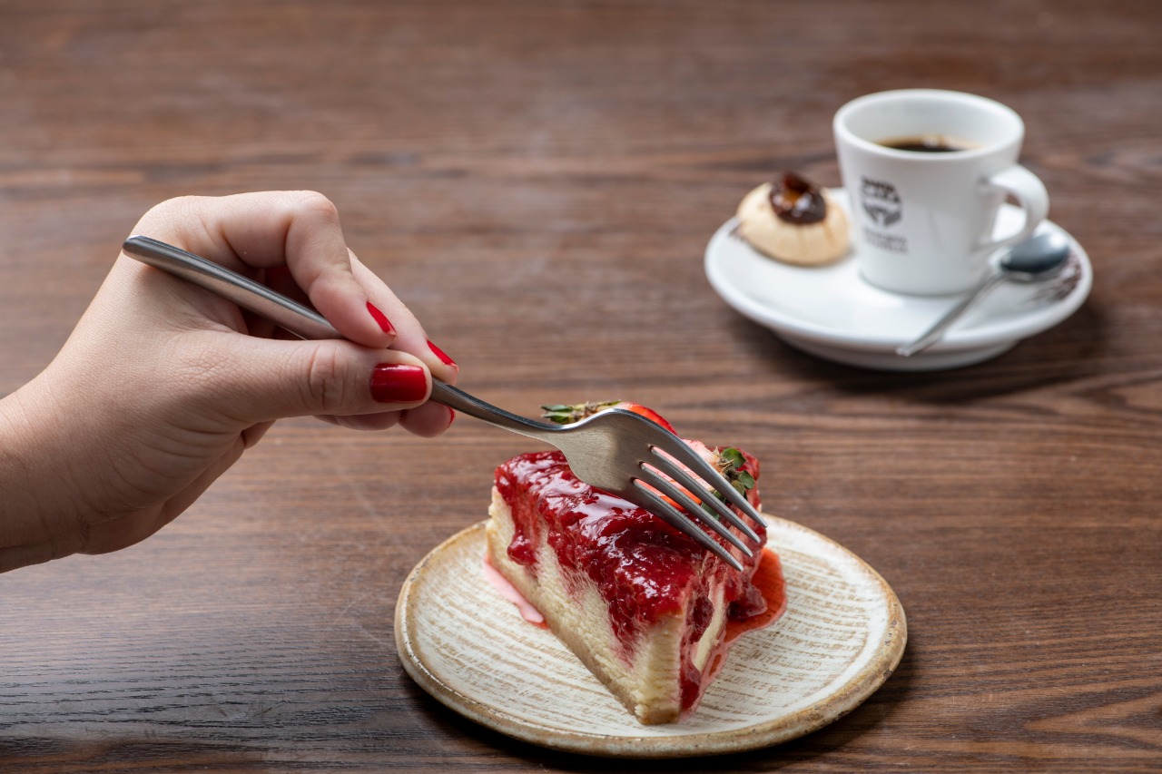 sobremesa: pessoa cortando um pedaço de cheescake de morango com uma xícara de café do lado