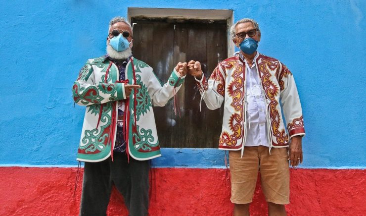 Cariri: na imagem, Ronaldo Fraga e Espedito Seleiro se cumprimentam com os punhos e posam para a foto. Ambos trajam vestes com as estampas feitas em couro tradicionais de Espedito, usam óculos e máscaras de proteção. Eles estão em frente a uma casa azul e vermelha.