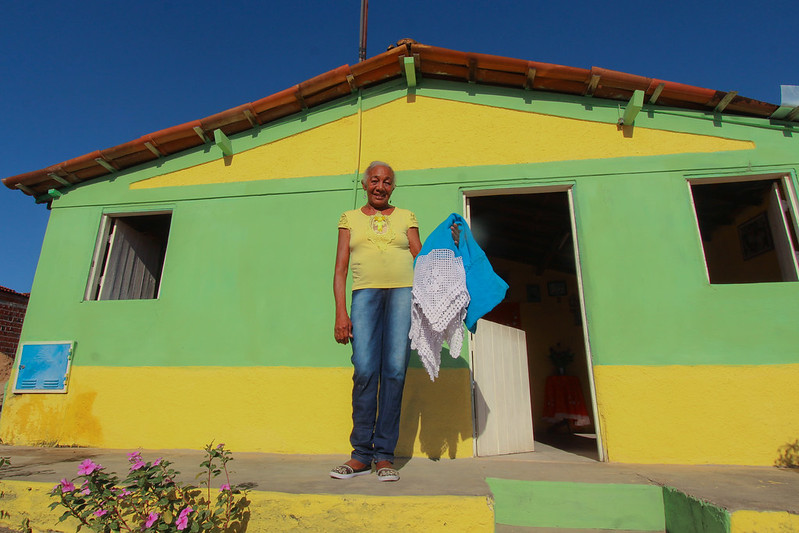 Museus orgânicos: uma mulher idosa vestida de camiseta amarela e jeans posa em frente a uma casa amarela e verde. Ela segura um tecido azul com renda branca e sorri para a foto.