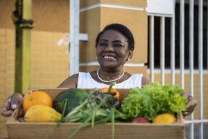 Mesa Brasil: uma mulher negra com cabelo preto e curto segura uma caixa com hortaliças e sorri para a foto. Ela usa colar e brincos de pérola e uma camisa sem mangas branca.