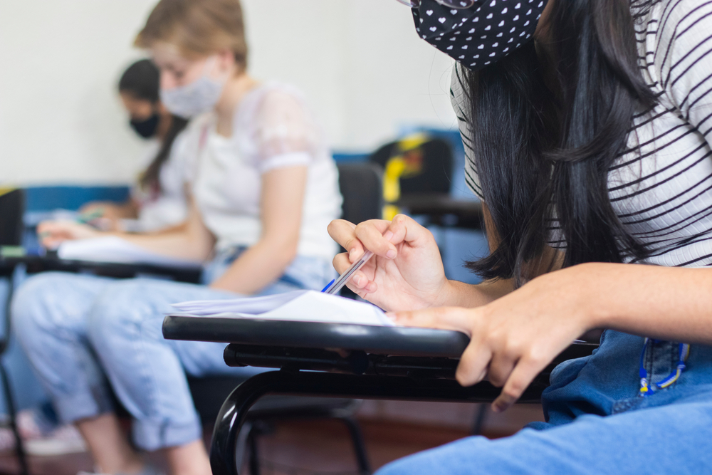 Curso pré-universitário: alunos de máscara, camiseta e calça jeans estão sentados em carteiras fazendo anotações.