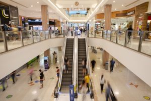 Dia do Comerciante: a imagem mostra a área interna de um shopping, com duas escadas rolantes e muitos clientes andando pelo espaço.