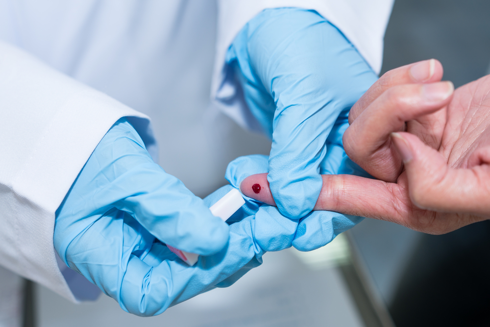 Hepatites virais: um par de mãos com luvas de proteção azuis aplica um teste no dedo de uma pessoa. Na ponta do dedo, há uma gota de sangue.