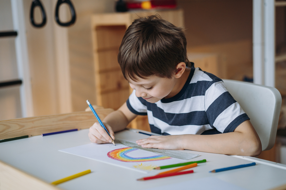 Contraturno no Criar Sesc: um menino branco e de cabelos castanhos desenha um arco-íris em uma mesa branca. Ele usa camisa listrada e segura um lápis azul.