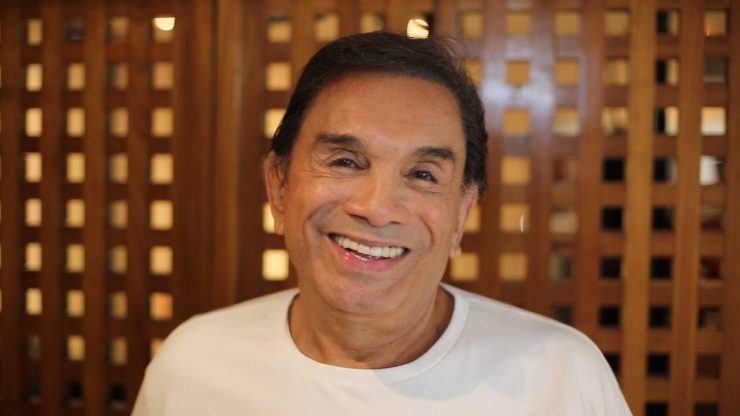 Dedé Santana sorri para a câmera. Ele está em frente a uma porta de madeira, tem cabelos pretos e cursos e usa uma camisa branca.