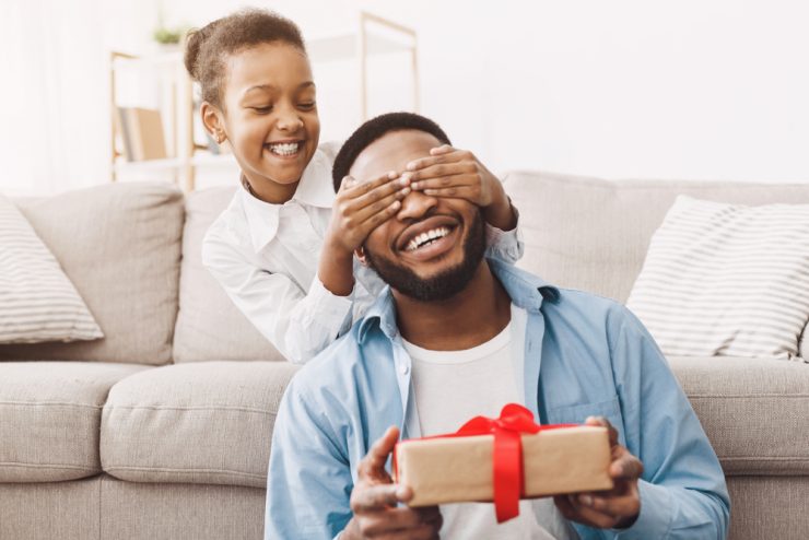 Dia dos pais: uma menina sorri enquanto cobre os olhos do pai com as mãos. Ele segura um presente com um laço vermelho. Ambos são negros, usam roupas claras e estão muito sorridentes. 