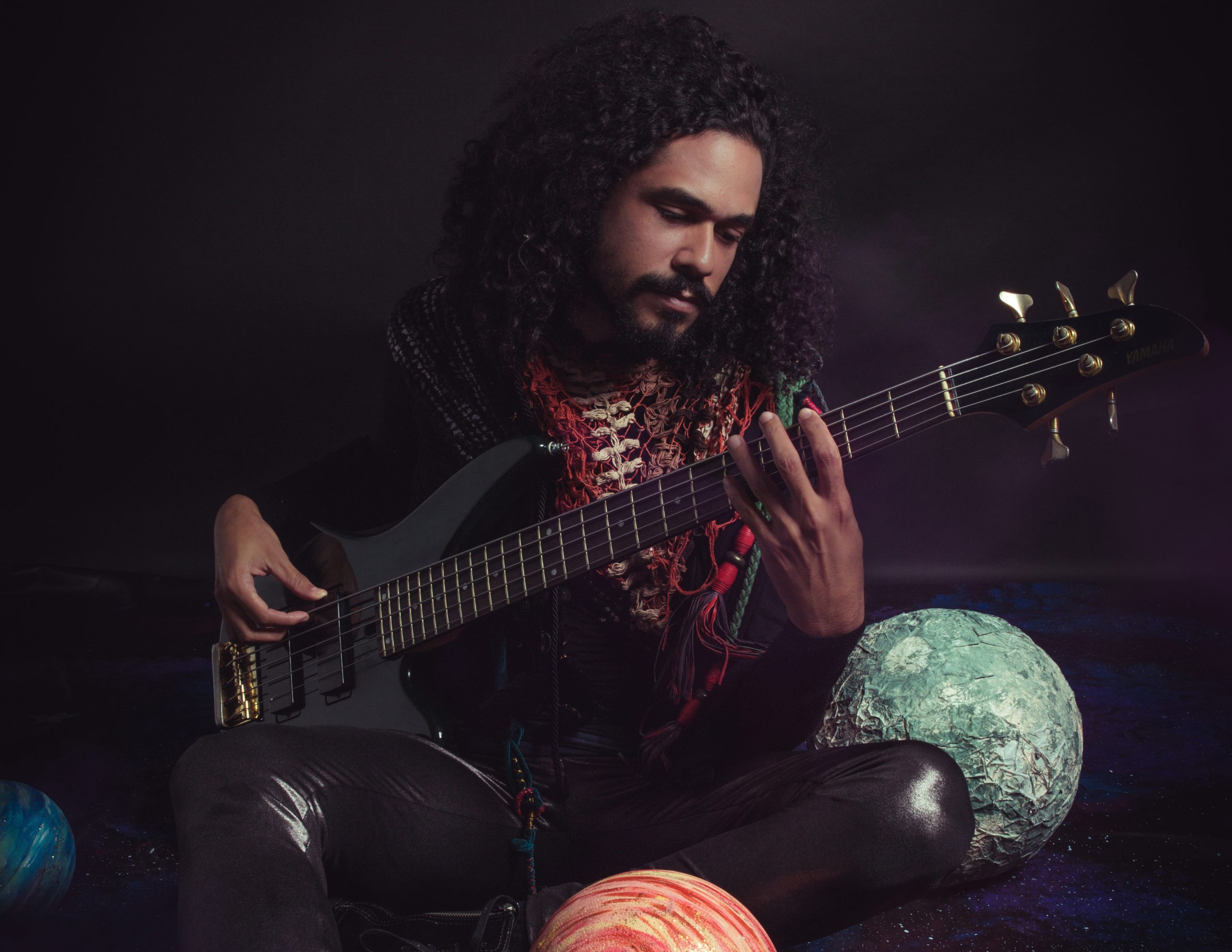Oficina de música: o músico Zéis segura uma guitarra em meio a um cenário com temática de planetas. Ele é um homem negro, usa cabelo grande e cacheado e bigode e um xale colorido.