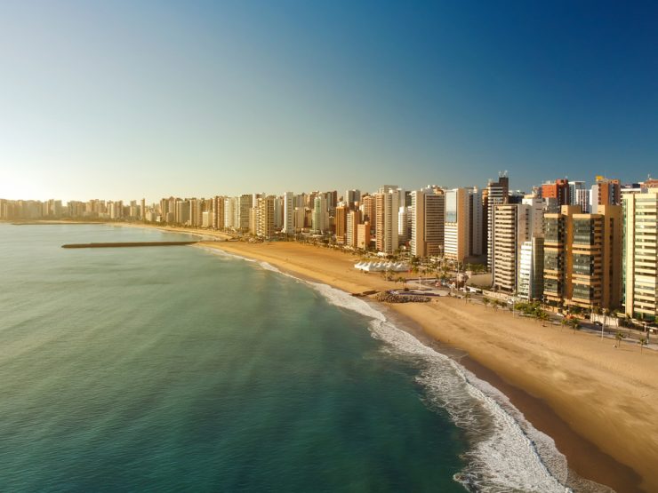 Dia Mundial do Turismo: imagem aérea da Praia de Iracema, em Fortaleza, em um dia de sol. Há prédios na orla, além de uma extensa faixa de areia.