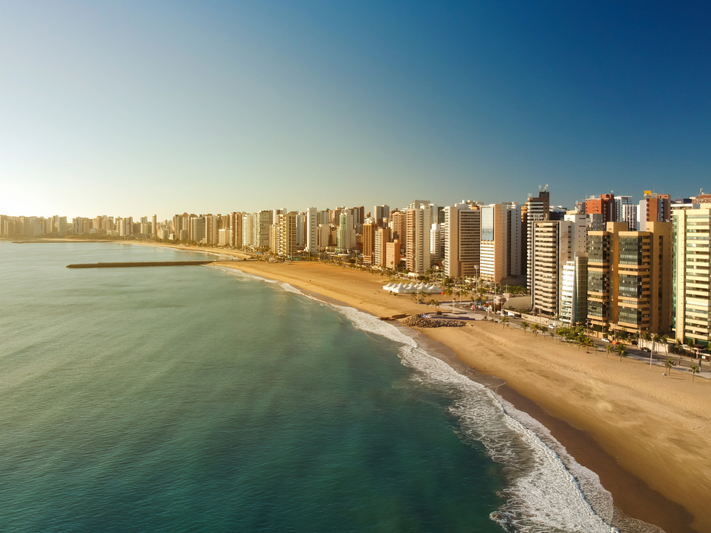 Dia Mundial do Turismo: imagem aérea da Praia de Iracema, em Fortaleza, em um dia de sol. Há prédios na orla, além de uma extensa faixa de areia.