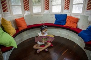 Espaço Mais Infância: uma menina de máscara de proteção, camiseta branca e short colorido lê um livro sentada no chão. Ao redor dela, há várias almofadas coloridas.