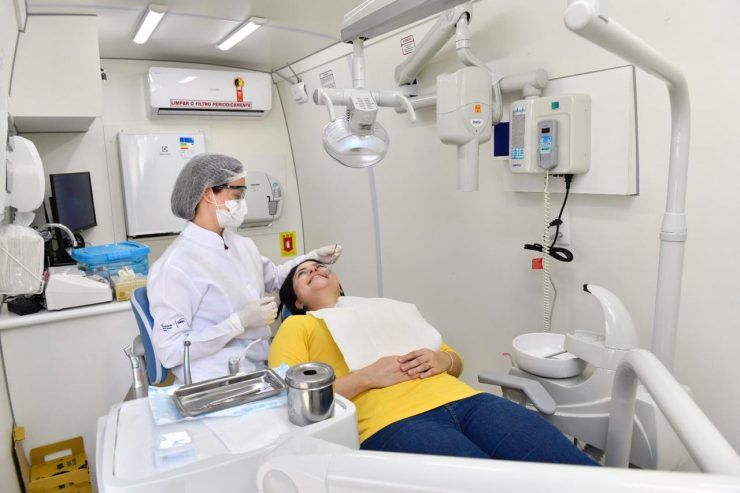 Dia do Comerciário: uma mulher branca de camisa amarela e calça jeans está deitada em uma cadeira num consultório de dentista. Uma dentista paramentada com jaleco, máscara, touca e luvas brancas examina sua boca.