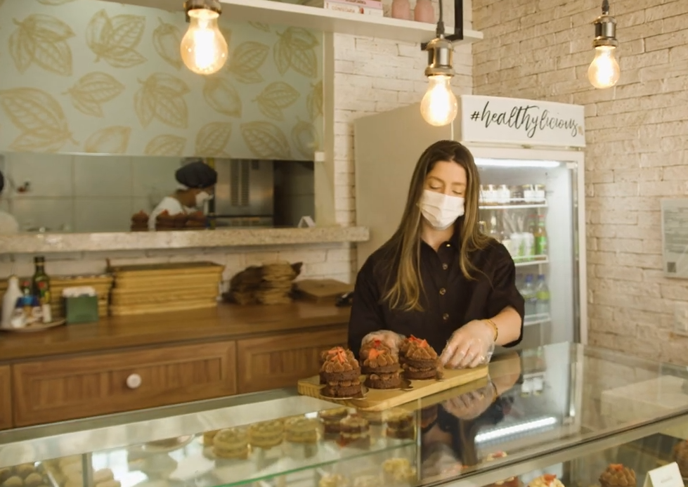 Cacao Saudável: foto da loja da Cacao Saudável. Uma mulher loira com máscara de tecido organiza doces em um balcão.