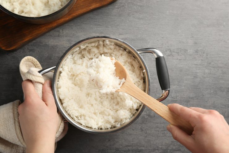 Bolinho de arroz: a imagem mostra uma panela cheia de arroz branco em cima de um balcão cinza e um par de mãos brancas. A mão direita segura um dos lados da panela e a esquerda segura uma colher de pau cheia de arroz.