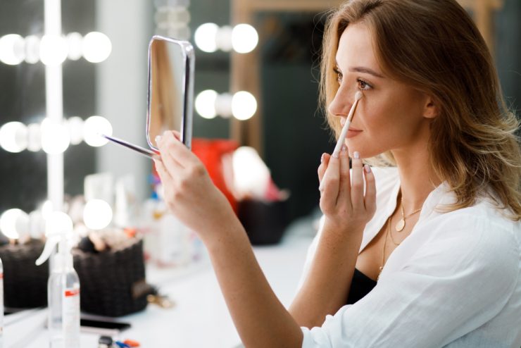 Automaquiagem: uma mulher branca, jovem e loira se maquia em frente a um espelho. Ela usa um pincel pequeno embaixo dos olhos e veste uma camisa branca