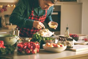 Receitas de Natal: com um avental vermelho quadriculado e uma camisa de flanela xadrez verde, um homem prepara uma receita de ave em uma mesa. Na imagem também há muitos ingredientes, como folhas, azeites e grãos.