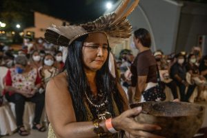 Herança Nativa: uma mulher indígena, com vestes tradicionais como cocar e colares, entrega uma cumbuca de barro para alguém. Ela está no foco da imagem, e há outras pessoas atrás dela.
