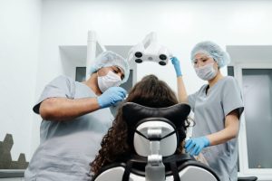 Mercado de saúde: duas pessoas com batas, máscaras e toucas cirúrgicas examina a boca de uma paciente.
